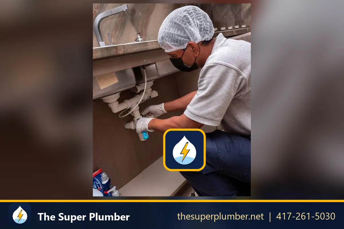 Worker in a hairnet and mask servicing the drain plumbing under a commercial kitchen sink - emergency plumber Newark