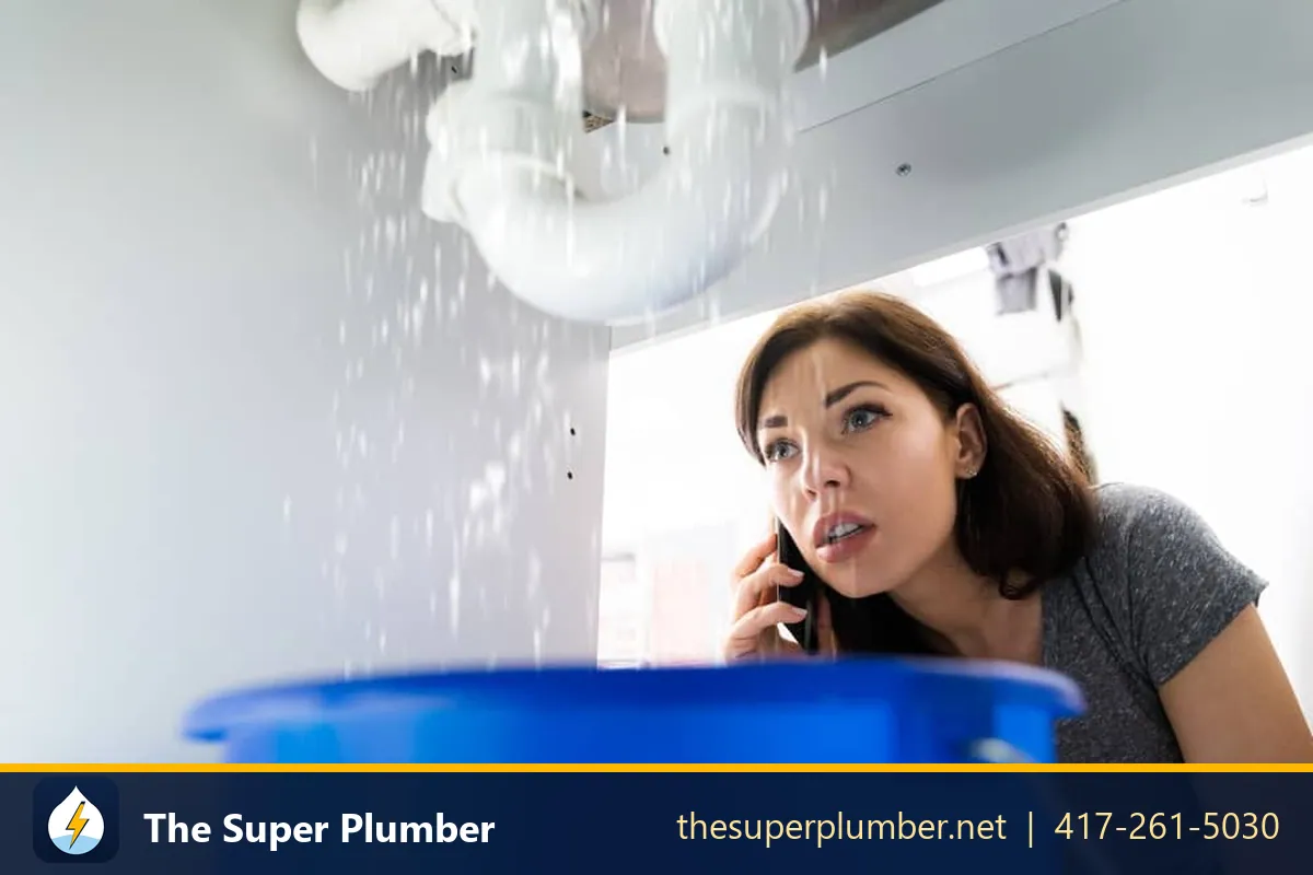 Woman phoning for help while water sprays from a pipe under a sink into a blue bucket - pipe breaks